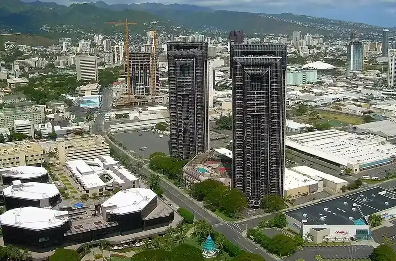 Two tall residential towers rising above low buildings in Honolulu, mountains behind
